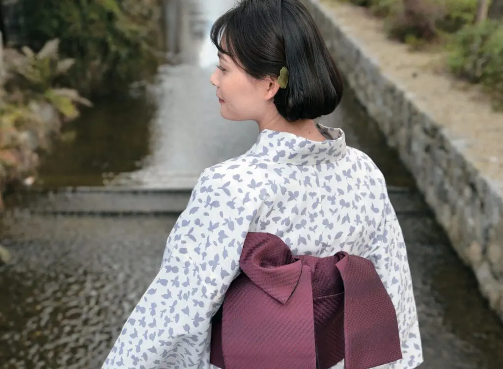 大きなワインレッド色の帯を締めた花柄の着物を着た女性が、静かな運河のそばに立っています。彼女は短い黒髪に小さな緑色のヘアピンを飾っており、カメラから背を向けて肩越しに見ています。背景には水、緑、石畳が描かれています。