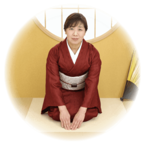 赤い着物と白と銀の帯を締めた女性が、伝統的な和室で膝をついて座っている。彼女は、木枠の丸い窓と、右側に立てられた扇子が描かれた黄色い仕切りの前に立っている。