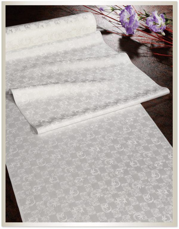 繊細で装飾的な花と曲線のデザインが施された、複雑な模様の白い布のロールが、暗い表面に部分的に広げられています。右側には、長い緑の茎を持つ紫と白の花がいくつか斜めに咲き、暗い背景に彩りを添えています。
