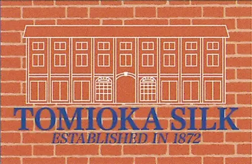 レンガ模様の背景に、複数の窓と中央のアーチを備えたレンガ造りの建物が白で縁取られたイラスト。建物のデザインの下には、青い文字で「TOMIOKA SILK」と書かれており、その下に「ESTABLISHED IN 1872」と添え字が書かれています。