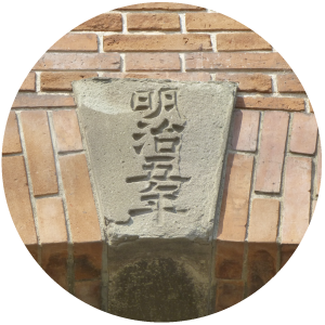コンクリートの銘板が埋め込まれたレンガの壁のクローズアップを示す円形の画像。銘板の表面には縦に並んだ日本語の文字が彫られています。レンガの壁には、赤みがかった色調とベージュ色の水平レンガと斜めのレンガが混在した模様があります。