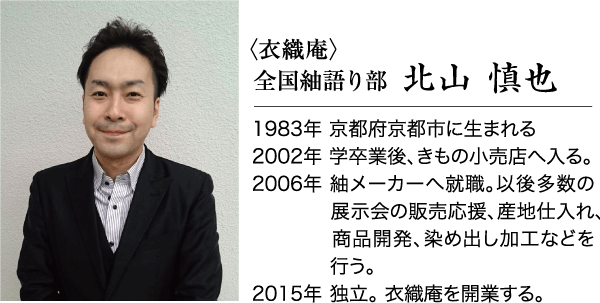 短い黒髪の男性が、薄い白のストライプが入ったライトグレーのドレスシャツの上に黒いスーツジャケットを着て、笑顔を浮かべている。彼は真っ白な壁の前に立ち、カメラに正面を向いて、両手を静かに前に置いている。