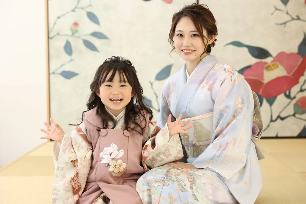 日本の伝統的な着物を着た女性と少女が、カメラに向かって温かく微笑んでいます。女性は花柄の淡いブルーの着物を着て、少女は七五三を祝うピンク色の着物を着ています。二人は畳の上に座っており、背景には花柄の衝立があります。スタッフBLOG