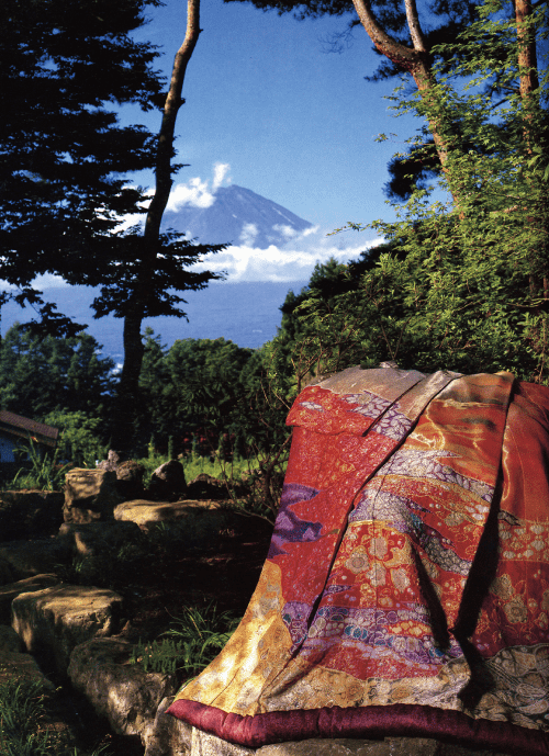 緑豊かな庭園の石のベンチに、赤、オレンジ、金色の模様が入った鮮やかな着物が掛けられています。背の高い木々が、明るい青空の下、遠くの雪を頂いた山々の眺めを縁取り、静かで絵のように美しい風景を作り出しています。.