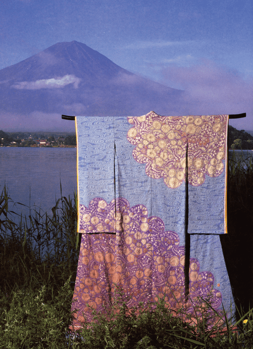 紫とオレンジの色合いの複雑な花柄が施された色鮮やかな着物が木製のラックに掛けられています。その着物は、澄んだ空の下の静かな湖と霧に包まれた雄大な山、そして青々とした緑の葦に囲まれた穏やかな背景を背に立っています。.