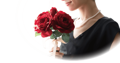 黒いドレスを着て真珠のネックレスをした女性が、緑の葉が付いた鮮やかな赤いバラの花束を持っています。背景は柔らかく照らされており、穏やかで穏やかな雰囲気が醸し出されています。画像は部分的に円形に切り取られており、花束と女性の穏やかな表情に焦点が当てられています。.