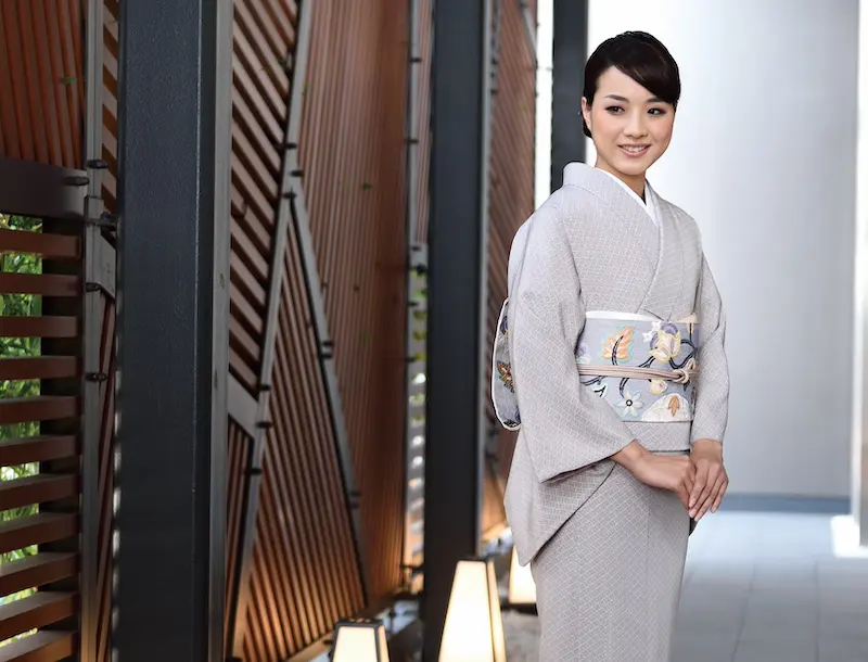 明るい灰色の着物に色とりどりの帯を締めた人物が、モダンな廊下に立っている。廊下の片側には木製のスラットが並び、照明器具からの柔らかな照明が暖かな雰囲気を醸し出している。人物は、これから起こるイベントに備えているかのように、優しく微笑み、優雅に手を組んで立っている。.