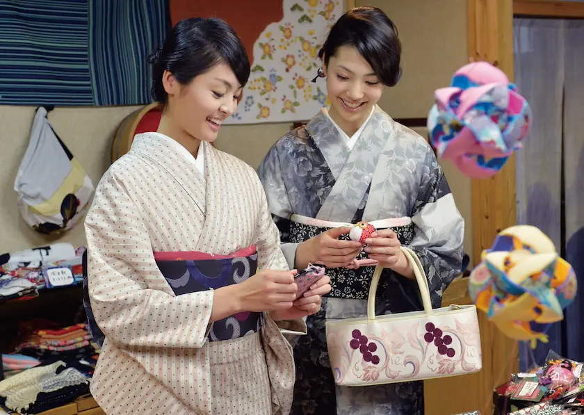 柄物の着物を着た2人の女性が居心地の良い店内に立って、小さな布製品を眺めながら微笑んでいます。スタッフBLOGにふさわしい楽しいシーンです。1人は花柄の淡い色のバッグを持っています。温かみのある照明と木製の家具に囲まれ、防寒の必需品を準備しているようです。.