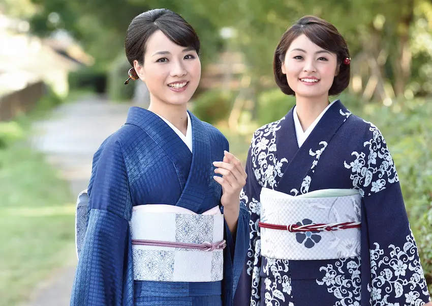 静かで緑に囲まれた小道で、2 人の女性が温かい笑顔を見せています。彼女たちは、複雑な模様の伝統的な青い着物と淡い色の帯を身にまとっています。穏やかな雰囲気を味わいながら、彼女たちの防寒対策は、スタイルと自然の思慮深い調和を物語っています。.