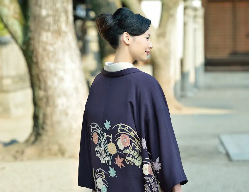 複雑な花と葉の模様が描かれた紫色の着物を着た女性が屋外に立って横を向いています。彼女の髪は上品なアップスタイルになっています。背景にはぼやけた木の幹と建物の壁があり、この穏やかな日本の風景の中で彼女が防寒の準備をしている様子が捉えられています。.