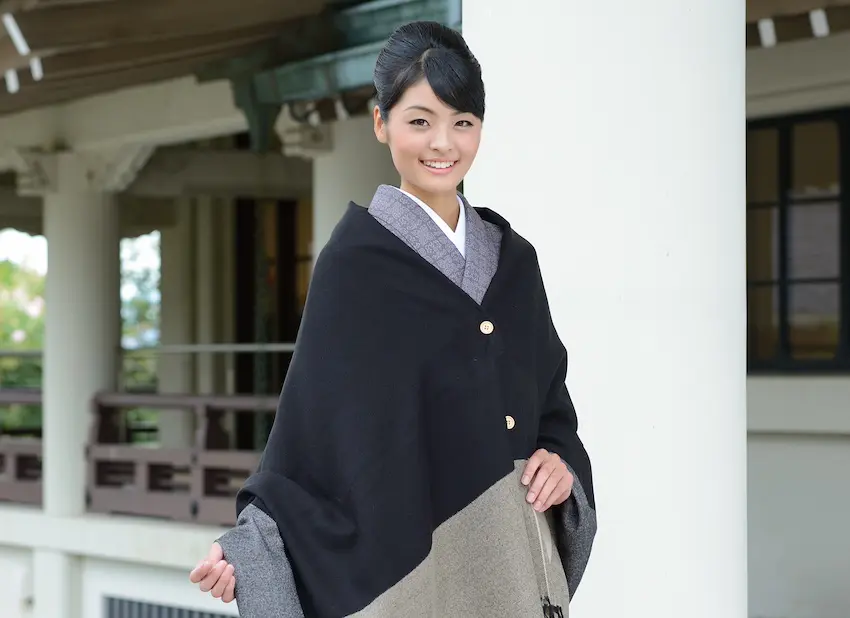 防寒を抱きながら、黒とグレーのショールを巻いた日本の伝統的な着物を着た女性が微笑んでいる。彼女は木造の建築物と柱の前に立ち、髪はきちんと整えられている。彼女の穏やかな態度は、ひんやりとした空気の中で暖かさを醸し出している。.