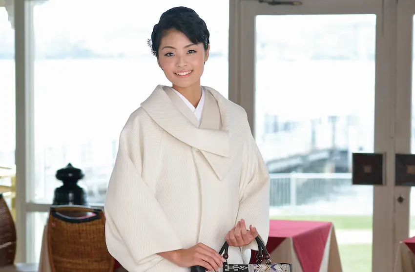 クリーム色の着物風の衣装を着た人が、柄物のバッグを持って屋内に立っています。黒髪を短く切り、笑っています。背景にはガラスのドアがあり、水と緑のぼんやりとした景色が見えます。中にはテーブルと椅子が置かれており、スタッフBLOGで防寒対策のヒントを読む訪問者を待っています。.