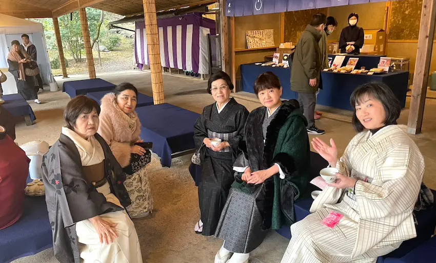 伝統的な着物を着た 5 人の女性が屋外のベンチに座っています。彼女たちは低いテーブルの周りに集まり、中にはティーカップを持っている人もいます。背景には小さなスタンドとその後ろに人がいて、紫と白の縞模様のカーテンがかかっています。雰囲気はくつろいでいて社交的です。.