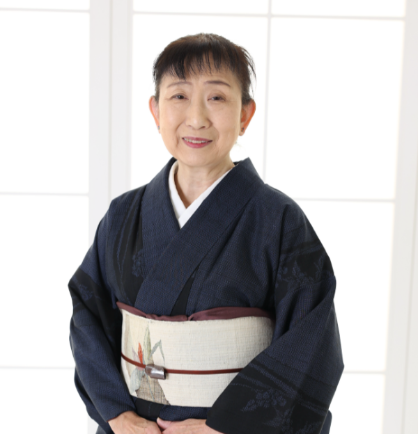 年配の女性が微笑んでいる。淡い色の細かな模様の着物に、小さな模様の入った淡い色の帯を締めている。髪はきちんと後ろに束ねられており、障子が張られた柔らかな光が差し込む窓の前に立っている。.