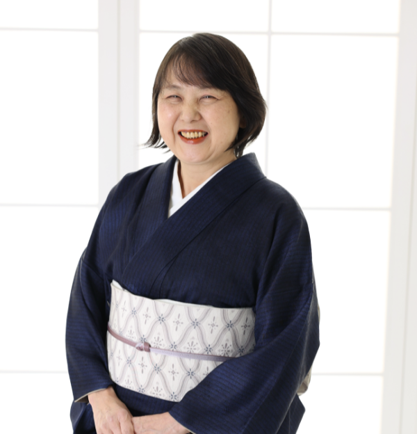 明るい窓の前に、淡い模様の帯を締めた濃紺の着物を着た女性が微笑みながら立っています。背景は柔らかく照らされ、伝統的な衣装が際立っています。彼女は短い黒髪で、両手を優しく前で組んでいて、楽しそうに見えます。.