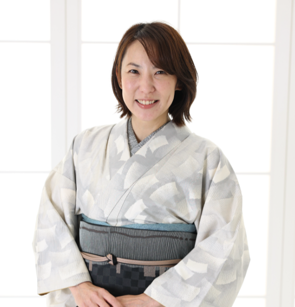 明るい逆光に照らされた窓の前に立って微笑む人物。淡い色の繊細な模様の着物と、濃い色の市松模様の帯を締めている。背景は柔らかく照らされ、人物の周りに穏やかな輝きを放っている。.