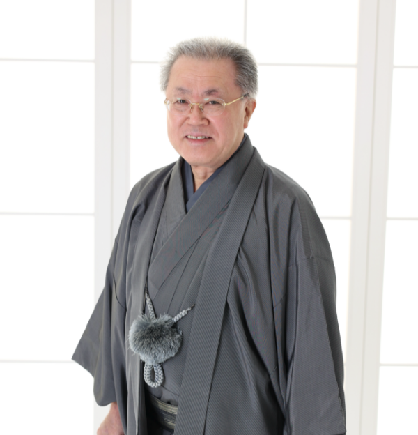 腰に飾り紐を巻いた伝統的な灰色の着物を着た年配の男性が微笑みながら立っている。彼は灰色の短い髪と眼鏡をかけている。背景には白い格子状のパネルを通して明るく拡散した光が映っている。.