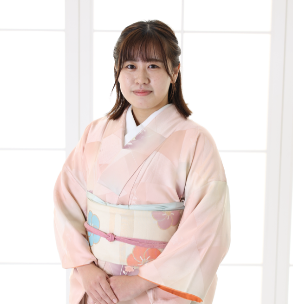 淡いピンク色の花柄の着物と華やかな帯を身に着けた人物が、室内の窓の前に立っています。背景は、格子模様の白い窓から差し込む柔らかな光で明るくなっています。.