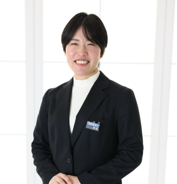 黒いブレザーと白いタートルネックを着て、笑っている短い黒髪の人物。ブレザーには名札が付いています。背景は白いパネルで柔らかく照らされています。.