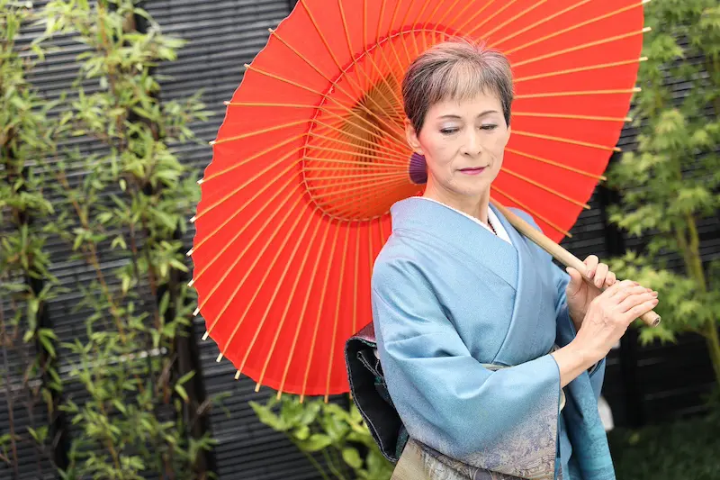 青い着物を着た人物が、緑の竹と暗い衝立を背景に赤い紙の傘を差しています。人物は穏やかな表情で下を向いています。.