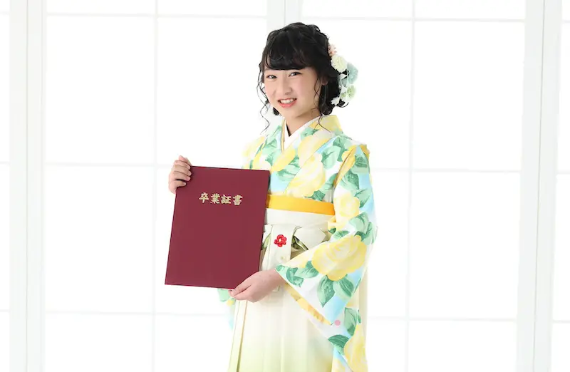 花柄の着物を着た人が、金色の文字が入った赤い卒業証書カバーを持っています。着物には明るい背景に黄色いバラと緑の葉が描かれています。彼らは黒い巻き毛で、白い花で飾られています。彼らの後ろの大きな窓からは、柔らかな白い光が差し込んでいます。.