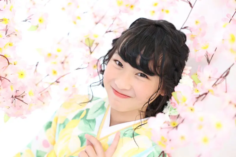 黒い巻き毛の人物が、ピンク色の桜に囲まれて微笑んでいます。黄色と緑の模様が入ったカラフルな着物を着ています。明るくソフトフォーカスされた背景が花のテーマを引き立て、穏やかで明るい雰囲気を醸し出しています。.