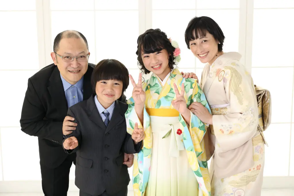 明るい窓の前で4人家族が笑顔を見せている。父親は眼鏡をかけ、黒いスーツを着ている。母親は淡い花柄の着物を着ている。色鮮やかな着物を着た娘とスーツを着た息子は、2人ともピースサインをしている。みんな楽しそうで仲良しそうに見える。.