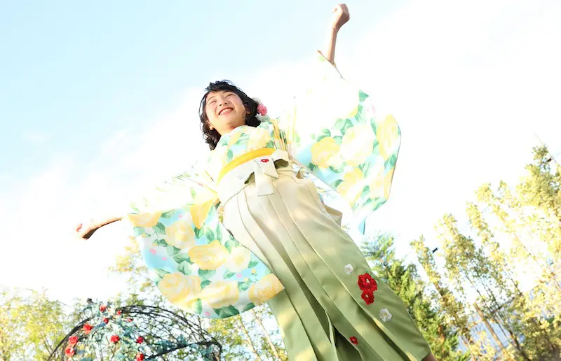 明るい青空を背景に、薄緑と黄色の花柄の着物を着た人が楽しそうに両腕を伸ばしています。着物には大きな黄色の花と赤い帯が描かれています。背景には太陽の光を浴びた緑の葉の木々が見えます。.