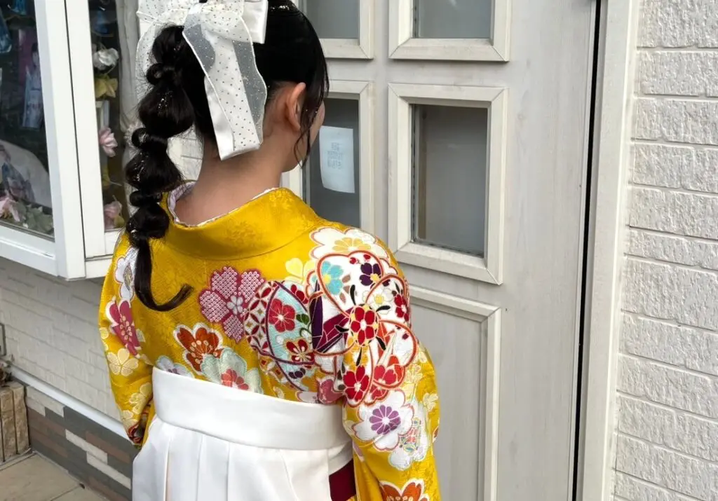 後ろから見た白いドアの前に立っている人物は、色とりどりの花柄が描かれた鮮やかな黄色の着物と白い帯を身に着けている。髪型は三つ編みで、水玉模様の大きな白いリボンで飾られている。左側には布製の花が飾られた店先の一部が見える。.