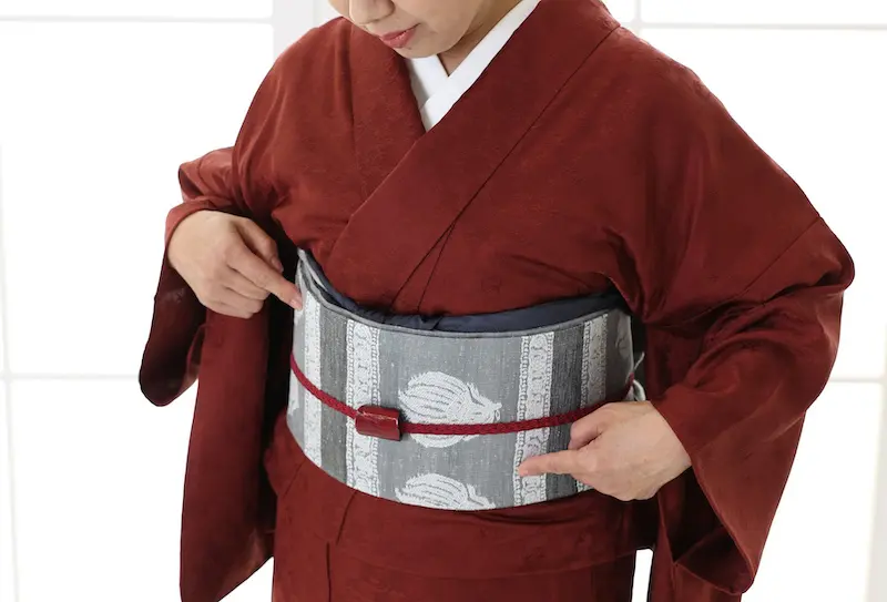 深紅の着物をまとった人物が、幅広の白とグレーの模様の帯を締め、細い赤いネクタイで締めている。明るい背景に上半身と手元が映え、伝統的な衿、長袖、草履が洗練された装いを完成させている。.