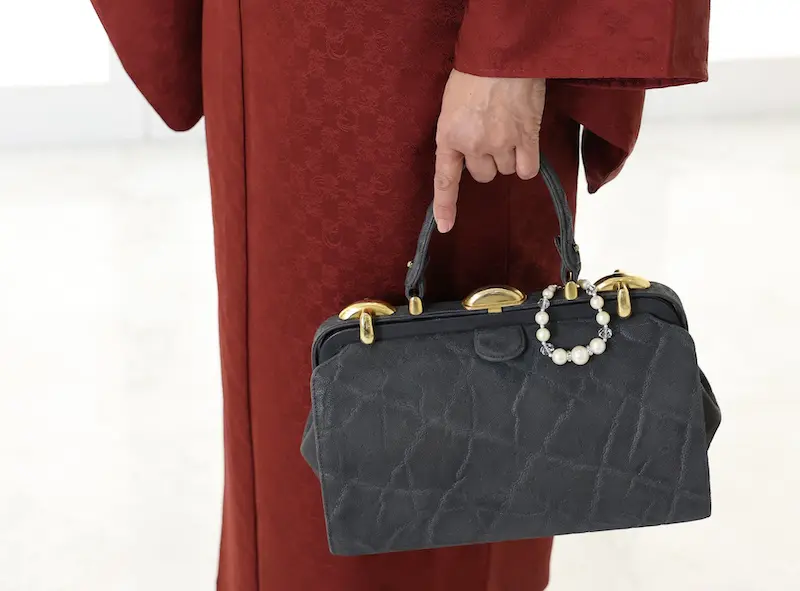 赤い模様の着物を着た人物が、金色の留め具と真珠の装飾が施された質感のある黒いバックを持っています。背景は明るく、ぼかされているため、伝統的な衣装と上品なアクセサリーが際立っています。.