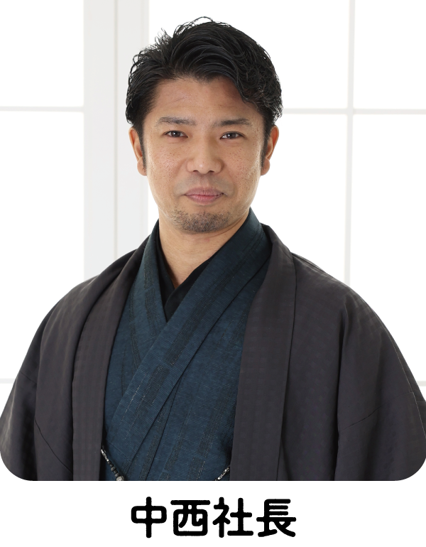 短くウェーブした黒髪に、整えられた髭を生やした男性が、紺色の伝統的な着物と黒い羽織を身に着けている。大きな窓の前に立ち、柔らかな笑みを浮かべ、自然光が顔を照らしている。.