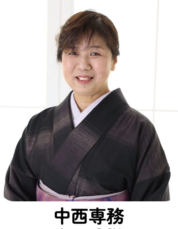 黒と紫の着物を着た黒髪のショートヘアの中年女性が、優しく微笑んでいる。彼女は白い枠の入った明るい窓の前に立っており、背景は柔らかな光に照らされている。.