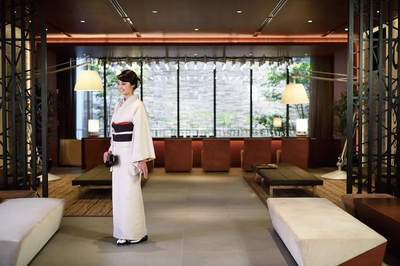 大きな窓から自然光が差し込み、ローテーブルとニュートラルカラーの座席が置かれたモダンでエレガントなラウンジで、伝統的な白い着物を着た女性が微笑んでいる。窓の外には緑と石垣が見える。.