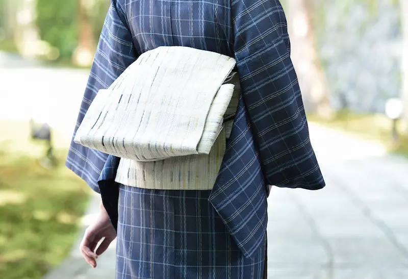 大きな淡い色の帯を後ろに締めた、伝統的な青い格子縞の着物を着た人物が、石畳の屋外に立っています。背景には緑の葉と太陽の光がぼやけています。写真は後ろから撮影されています。.