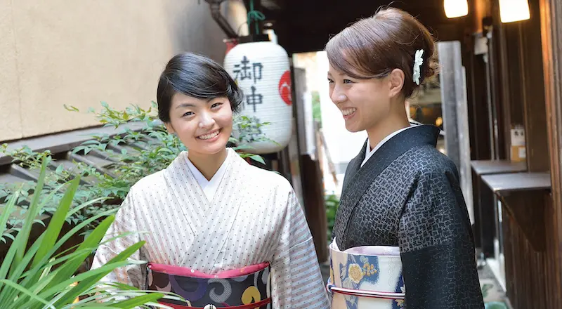 伝統的な着物を着た二人の女性が、狭い歩道に寄り添って微笑みながら立っている。周囲には緑が生い茂り、背景には木造の建物に取り付けられた、日本語の文字が書かれた白い提灯が吊るされている。.