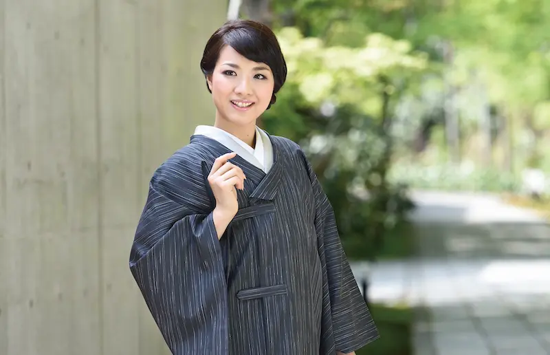 白い襦袢の上に濃い灰色の縞模様の着物を着た笑顔の女性が屋外に立っている。短い黒髪で、着物の衿の片側を押さえている。背景には緑、コンクリートの壁、そして舗装された小道が描かれている。.