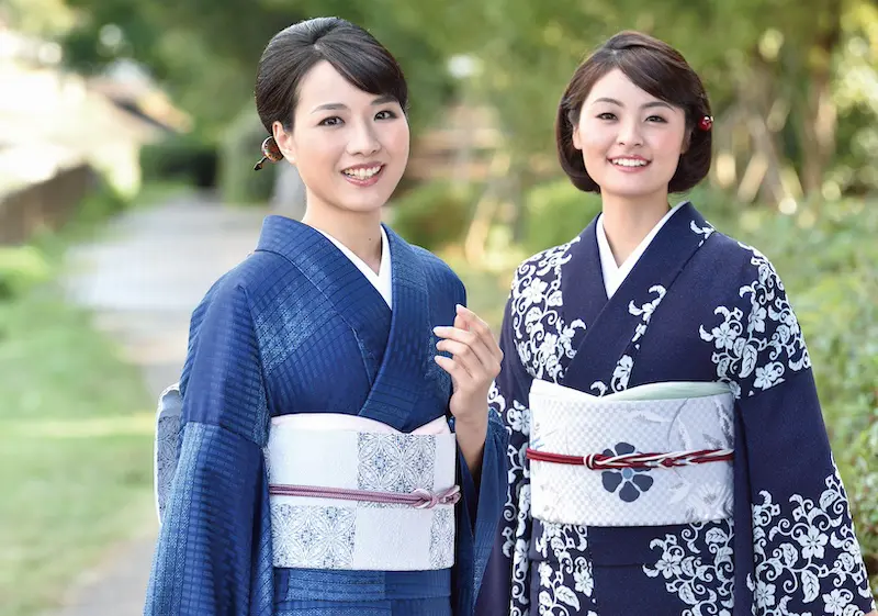 二人の女性が屋外に立って微笑んでいる。青と白の複雑な模様が描かれた日本の伝統的な着物を着て、二人とも黒髪をきちんとスタイリングし、日差しが差し込む小道の緑に囲まれている。.