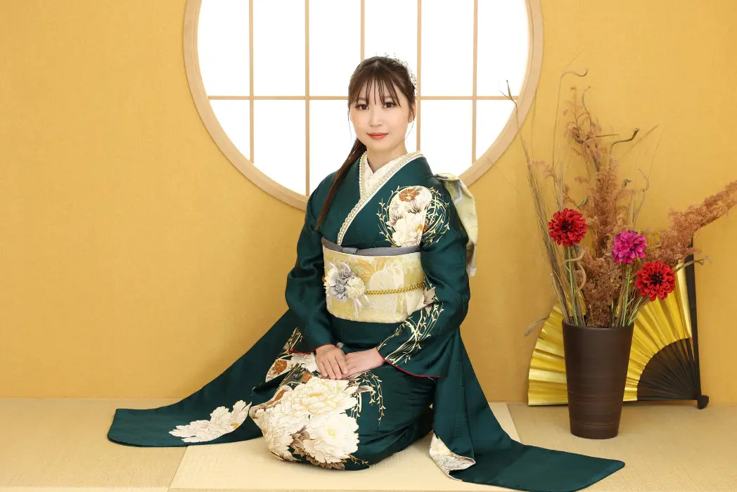 濃い緑色の花柄の着物を着た若い女性が、丸い窓の前で畳の上にひざまずいている。前髪のある茶色のストレートヘア。彼女の横には赤と紫の花を生けた背の高い花瓶と黄色い扇子が置かれ、すべて日焼けした壁を背にしている。.