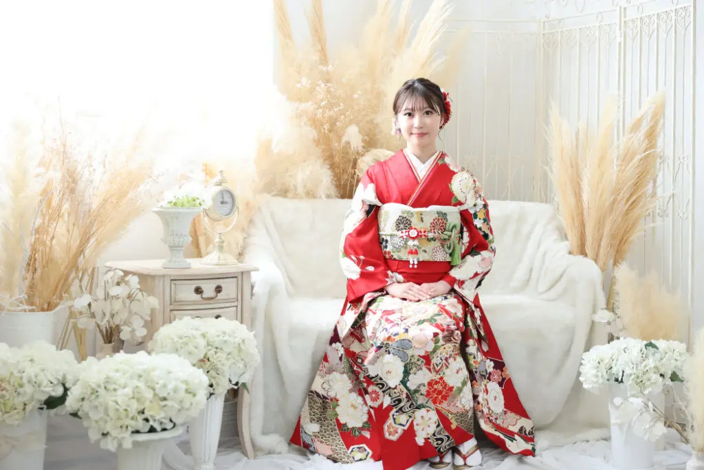 鮮やかな赤い花柄の着物を着た女性が、白いソファに優雅に腰掛け、膝に手を組んでいる。エレガントに飾られ、柔らかな照明に照らされた部屋の中で、白い花とパンパスグラスに囲まれた彼女は、静謐で伝統的な雰囲気を醸し出している。.