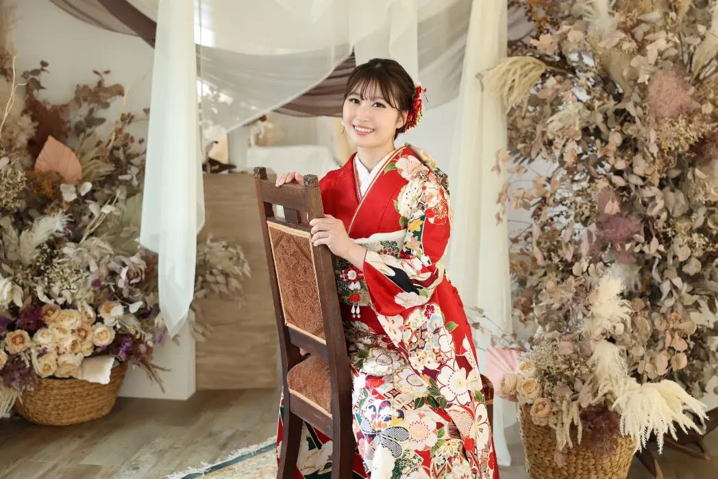 色とりどりの花柄の着物を着た笑顔の女性が、ドライフラワーやバスケット、柔らかなドレープで飾られた部屋の木製の椅子に後ろ向きに座っている。赤いヘアアクセサリーをつけた凝ったヘアスタイルで、カメラに向かって自信たっぷりにポーズをとっている。.