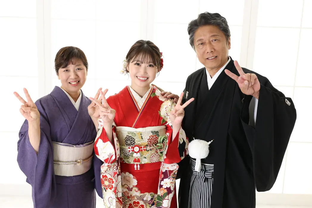 日本の伝統的な衣装を着て微笑みながら立っている3人。中央の若い女性は赤い花柄の着物を着て髪飾りをつけている。左の女性は紫の着物を、右の男性は黒い着物を着ている。全員がピースサインを掲げている。.
