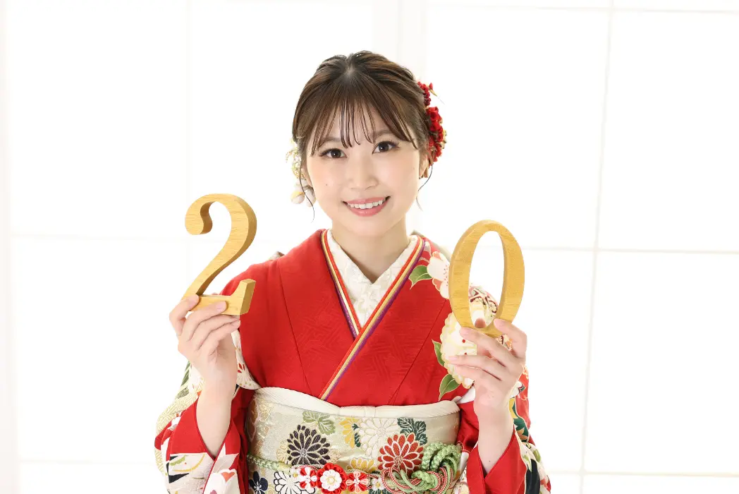 カラフルな赤い着物を着た若い女性が、20を意味する「2」と「0」の大きな木製の数字を持って微笑んでいる。髪には花の飾りをつけている。彼女は真っ白な窓の前に立ち、特別な日を祝っている。.