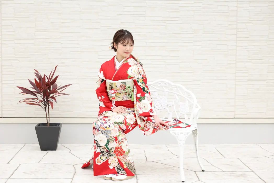 鮮やかな赤い花柄の着物を着た若い女性が、白い金属製のベンチに優雅に座っている。髪に花を飾り、着物の袖を押さえて下を向いている。彼女の左側には赤い葉をつけた鉢植えが、淡い石壁を背にして置かれている。.