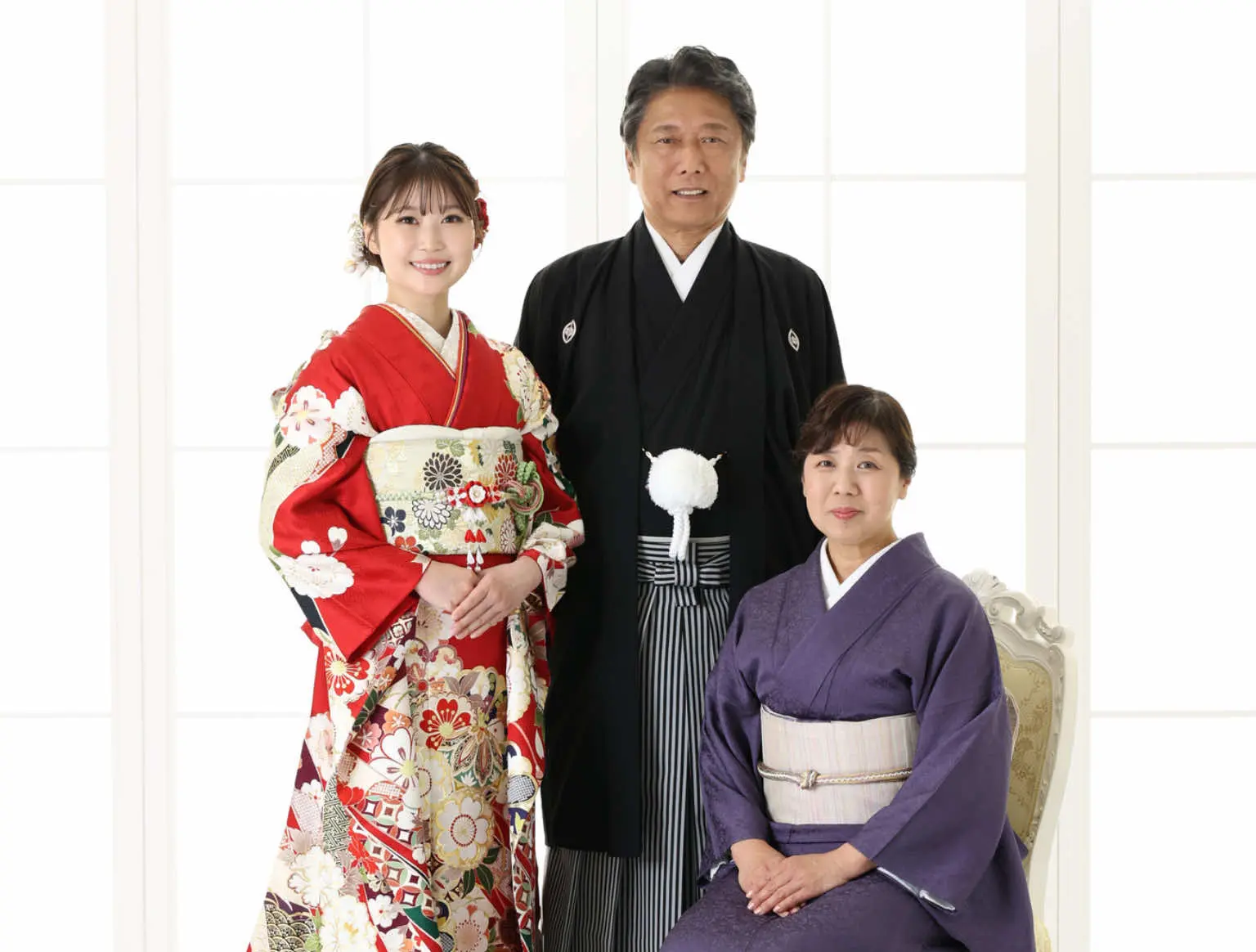黒と白の伝統的な和服を着た年配の男性の隣に、赤い花柄の着物を着た若い女性が微笑みながら立っている。紫色の着物を着た年配の女性が椅子に座っている。3人とも背の高い明るい窓の前でポーズをとっている。.