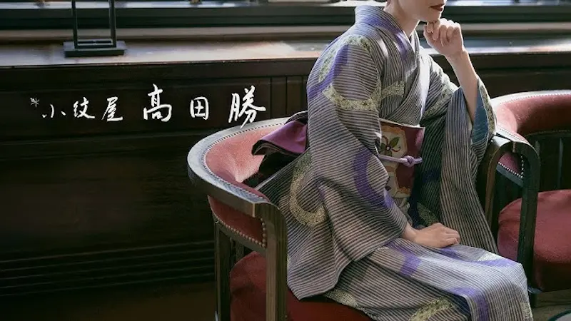 縞と花柄の着物に紫の帯を締めた人物が、赤いクッションの椅子に優雅に座り、時代とユニークな着方に思いを馳せている。画像の左側には日本の書道が見える。.