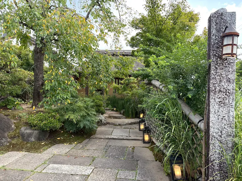灯籠が並ぶ石畳の小道は、緑豊かな木々の間を通り、伝統的な木造の日本建築へと続く。風化した石柱と竹垣が、着物の散歩やもみじ狩りに最適な落ち着いた庭の風景をさらに引き立てている。.