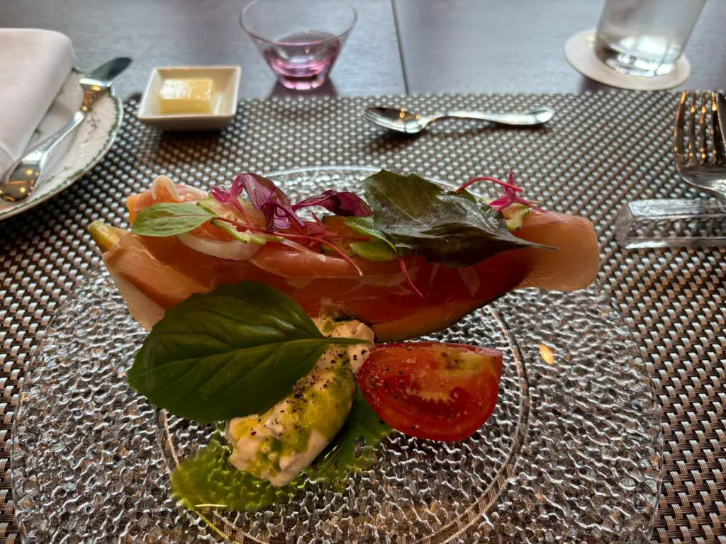 透明なガラスの皿の上には、生ハムのスライスとフレッシュグリーン、マイクログリーン、バジルがのっている。その横には、半分に切ったプチトマト、クリーミーなサラダ、グリーンソースが添えられている。.