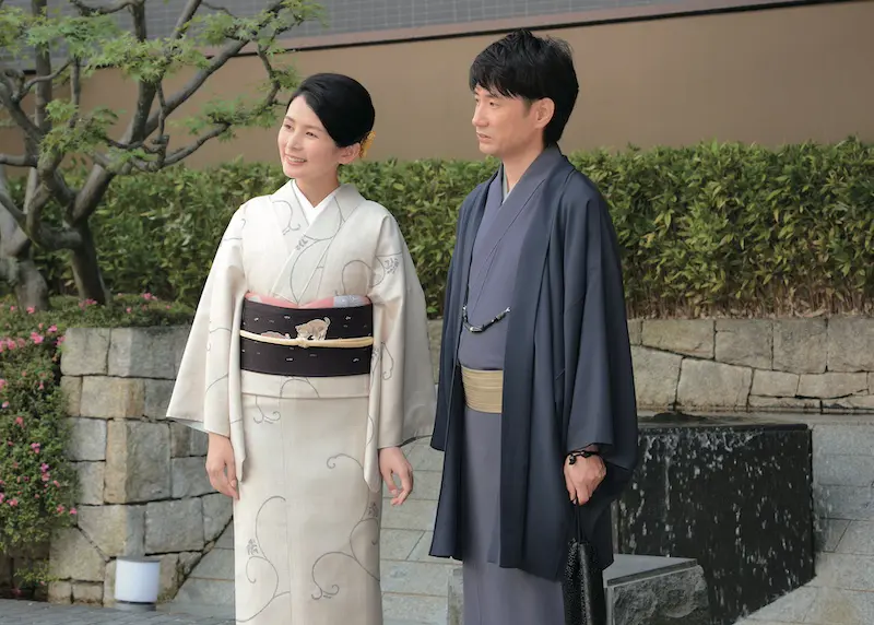 石垣と緑の前で、エレガントな着物姿の女性と男性。ふたりの笑顔が新春のスピリットを表現している。.