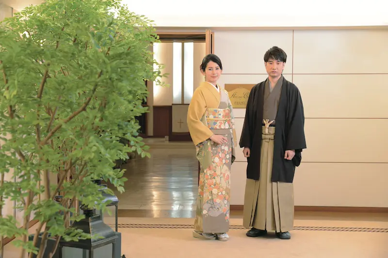 室内で大きな観葉植物のそばに立つ女性と男性。エレガントな新春着物-色とりどりの花柄の着物に帯、フォーマルな黒の羽織にベージュの袴。温かみのある照明と木製の装飾が、落ち着いた上品な雰囲気を醸し出している。.